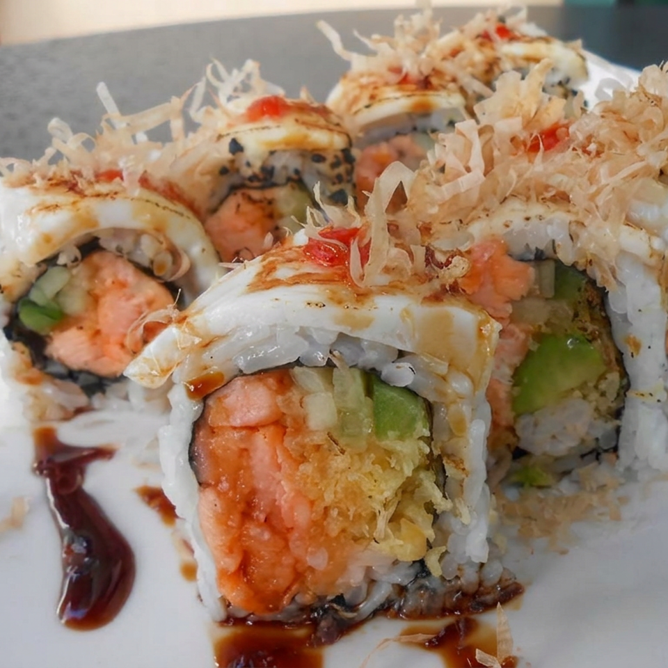 Close-up of sushi rolls topped with shredded garnish, salmon, avocado, and drizzled sauce on a white plate.